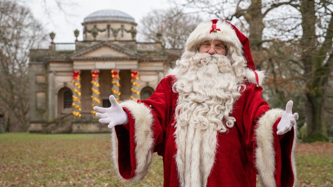 Father Christmas in traditional red clothes and a big white beard in front of a Chapel covered in Christmas lights.